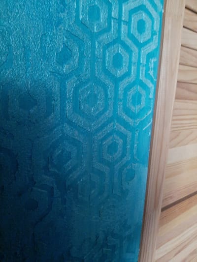 The image shows a close-up, eye-level view of a textured turquoise wall with a hexagonal pattern, possibly wallpaper or a painted finish. The texture appears somewhat distressed, with areas of lighter and darker shades indicating depth and wear. To the right of the wall, a wooden frame borders the scene, and beyond that, a surface of light-colored wooden planks is visible, arranged horizontally. The scene is indoors, with the wooden planks suggesting it might be part of a room or furniture. There are no people or animals present. The lighting suggests an indoor environment, possibly with artificial light. There is no information within the image to determine its location in Elizavetino, Russia.