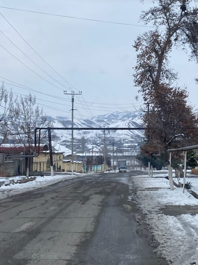 This image displays a street scene in Norayburi, Uzbekistan, under an overcast winter sky. The view is directed down an asphalt road, which appears wet and slushy with visible tire tracks, flanked by accumulated dirty snow and slush on both verges.
In the midground, large, dark-colored utility pipes are elevated horizontally across the frame on the left. Behind these pipes, a tall utility pole supports multiple cross arms and numerous overhead power lines that extend across the upper portion of the scene. To the left of the road, behind the pipes, are buildings including a low, light-colored structure with rectangular features and a grey concrete block wall, alongside several bare deciduous trees.
On the right side of the road, a large deciduous tree retains clusters of dry, brown leaves, partially obscuring smaller trees and additional utility poles.
Further down the street in the central midground, a white vehicle, consistent with a van or small car, is discernible. Beyond this vehicle, a cluster of buildings with snow-covered roofs and a prominent elevated structural element, possibly industrial infrastructure or a bridge, are visible. The background is dominated by a range of snow-covered mountains under a pale, diffuse sky, with some areas showing exposed darker terrain. No individuals are explicitly visible in the scene.