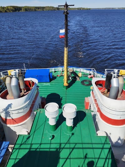 View from the deck of a ship on the Volga River in Russia. In the foreground is the green deck of the ship, with white and red equipment housings and two white vents. In the midground is a black mast with a Russian flag attached. In the background is the blue water of the Volga River, with a green tree line on the horizon. The sky is blue and cloudless.