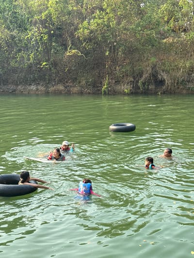 In Tambon Nam Phrae, Thailand, a daytime outdoor scene captures several individuals swimming in a body of water, likely a lake or river, bordered by dense vegetation on the left side of the frame.
Six individuals are visible in the water. Some appear to be children, and at least one is wearing a life vest. Two dark-colored inflatable inner tubes are present; one is unoccupied and floating, while another is being used by one or two of the swimmers. The water is green, suggesting the presence of algae or other organic matter. The vegetation on the bank is lush and dense with varying shades of green.