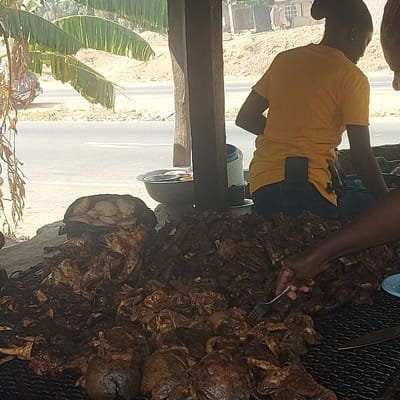 The image depicts a roadside food stall operating during daylight in Assai Sabongida, Nigeria. In the foreground, a substantial pile of dark, roasted or grilled meat, consistent with local cuisine, rests on a large metal grate. A dark-skinned hand, entering the frame from the right, holds a silver fork, selecting a piece from the meat pile. A blue and white plate and a knife are visible on the grate adjacent to the hand. To the left, behind the meat, a black bag contains a stack of light-colored, round food items.
In the midground, a person, viewed from the back, wears a yellow short-sleeved t-shirt and dark trousers, with a black smartphone secured in their rear waistband. This individual stands behind a wooden counter or stall support structure. Multiple metal bowls, some stacked, are positioned on the counter beneath the structure.
The background features a paved road with a visible white shoulder line. Beyond the road, a dry, sandy embankment rises. Green banana leaves are prominent on the far left, indicating tropical vegetation. A motorcycle is partially visible on the road in the distant left. Further in the background, across the embankment, several light-colored buildings with dark roofs are discernible, suggesting a settlement.