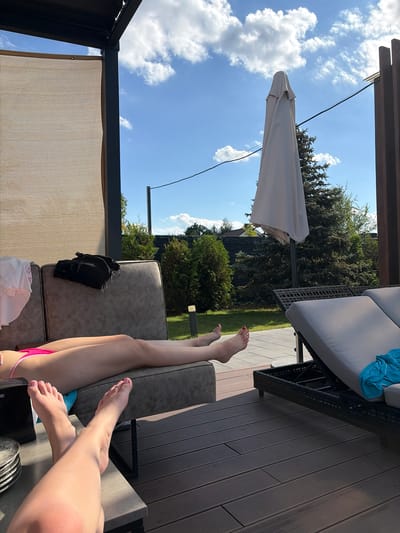 The image depicts a serene outdoor scene in Slobozhanske, Ukraine, featuring two individuals relaxing on a patio. In the foreground, one person's legs are visible, resting on a table. Their feet, adorned with nail polish, are casually positioned. Behind them, another individual lies on a lounge chair, their legs extended. The background reveals a well-maintained lawn with lush green trees and a covered seating area, likely part of a residential property. A closed umbrella stands nearby, suggesting a break from the sun. The sky above is a vibrant blue, dotted with fluffy white clouds, indicating pleasant weather. The overall atmosphere is one of relaxation and leisure in a suburban setting.