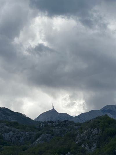 The image shows a mountain peak in Očinići, Montenegro, partially obscured by clouds. A transmission tower is visible at the summit. The mountain's slope is covered in vegetation. The sky is overcast with grey clouds.