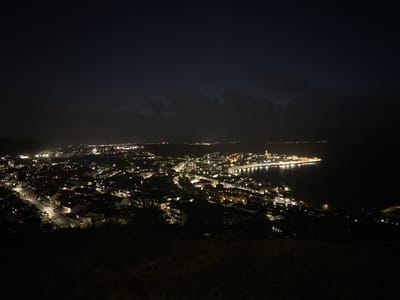 The image shows a nighttime view of Gourock, United Kingdom from an elevated perspective. The town’s lights illuminate the landscape, with residential areas and streets visible. The coastline is defined by the presence of water, and a pier or promenade juts out into the sea. There is no indication of any specific person, object, or event of interest. The foreground is a dark, elevated terrain that is overgrown, obscuring the details of the ground below.