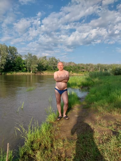 An adult male stands on the bank of a river, wearing blue swim briefs and black sandals. He has his arms crossed and is facing the camera with a neutral expression. The river is calm and reflects the blue sky with scattered white clouds. Lush green vegetation lines the opposite bank, with trees forming a dense backdrop. The foreground consists of a muddy bank with grass. The image appears to be taken outdoors during the daytime, likely in a rural or natural setting. The location is identified as Chkalov, Russia.