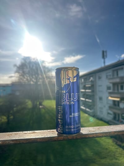 A blue Red Bull "The Blue Edition" energy drink can, featuring "Heidelbeer" (blueberry) flavor and the German slogan "Belebt Geist und Körper," stands upright on a rusted metal railing. The scene is set outdoors from an elevated perspective in Höri, Switzerland. The upper-left quadrant of the frame is dominated by a bright sun, causing a significant lens flare. The sky is clear blue with scattered white clouds. In the background to the left, a green grassy area and a tree with bare branches are visible, along with a building featuring blue elements. To the right, a multi-story residential building with multiple balconies and light-colored awnings is present. A cellular communication tower is discernible on a rooftop in the far distance to the right. No individuals, explicit actions, or interactions are depicted.