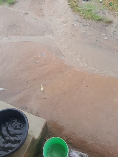 This image depicts an outdoor ground-level scene in Ifo, Nigeria, primarily characterized by flowing water and two water collection vessels. The foreground features a light-colored concrete slab extending horizontally, upon which a black, round plastic basin is positioned in the lower-left quadrant, partially filled with clear water exhibiting ripples. A bright green plastic bucket, also containing clear water, is visible in the lower-right corner, adjacent to the concrete slab.
Beyond the concrete slab, the terrain consists of reddish-brown, wet, sandy earth. A channel of turbid, brownish water actively flows from the upper right toward the lower-left, displaying visible ripples and current. The ground surrounding the flowing water is uneven and littered with various debris, including crumpled light-colored plastic wrappers, a colorful wrapper, and a small square of patterned material. Sparse green grass, interspersed with additional refuse, lines the upper edge of the frame and the bank of the flowing water in the upper right quadrant. No individuals are present in the scene.