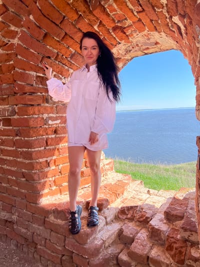 A woman stands inside a brick structure in Шуран, Russia. The structure has a weathered red brick interior and looks out onto a body of water with a grassy bank on the opposite side. The woman has long dark hair and is wearing a white shirt-dress and black sandals. The sky above is blue.