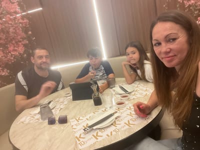 The photograph captures a family of four seated around a round table in a restaurant, possibly in the Torreforta, Spain, location of K10 Sushi, indicated by the branding on the table setting.

The family is positioned around the table with the woman taking a selfie, capturing herself and the rest of her family. To her left is her daughter, who is resting her head on her hand with a neutral expression. Next to her is a young boy drinking from a glass with a straw, and further to the left is a man, likely the father, with a hand raised and an ambiguous expression.

The table is adorned with sushi place settings including white plates, chopsticks, and small dishes containing a red-colored sauce. A tablet device is also on the table in front of the boy.

The restaurant is dimly lit with artificial lighting. A decorative floral display of pink blossoms is visible in the background to the left of the man, suggesting a themed or stylish ambiance. The background appears to be a soft beige color, and the walls are padded or upholstered.