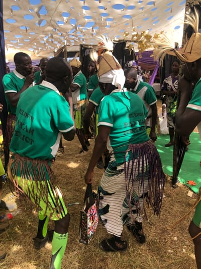 This media file captures a cultural event taking place outdoors in Kerang, Nigeria, under bright daylight. The scene features several individuals, predominantly men, dressed in distinctive attire.
The main subjects are a group of people, appearing to be performers or participants in a ceremony. They wear matching green polo shirts with white collars and sleeve trim. On the back of these shirts, text is visible, with one prominent example reading "BEL BWOH" and "BANYAM DISTRICT," and another partially showing "NYAM DISTRICT." Many of the men wear traditional headwear, including woven straw hats adorned with white plumes or white fabric wraps. Their lower attire consists of colorful, fringed wraps or skirts worn over trousers or directly on their legs; these fringes are visible in shades of purple, brown, and green. Some individuals also have green athletic socks. One person near the foreground holds a black object, possibly a bag or an instrument, while a colorful, patterned bag rests on the ground nearby. In the background, a woman wearing a patterned purple and black dress is partially visible.
The setting is an open-air area with dry grass and earth, suggesting a warm and sunny climate. A large, light-colored fabric structure with decorative circular cutouts provides shade on the left, possibly a tent or canopy. Behind the participants, hints of festive decorations can be seen, including purple and gold fabrics and some green ground cover. The overall impression is one of a vibrant community gathering or performance.