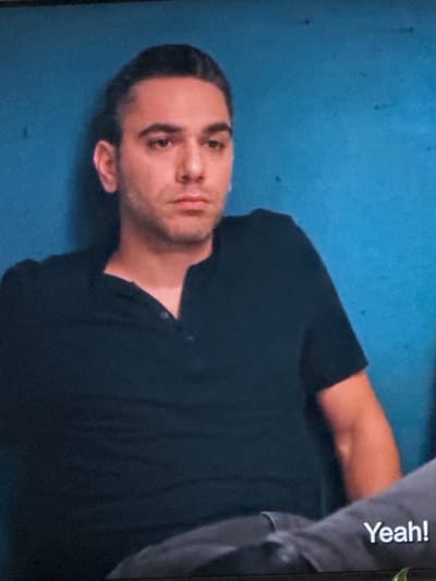 In this close-up shot of a man, he is leaning against a blue wall. He is wearing a black Henley shirt. His hair is dark and slicked back. His expression is neutral, and he is looking slightly off-camera. In the foreground, the word "Yeah!" is visible in white text on a grey surface, possibly a piece of clothing or furniture. The image is grainy, suggesting it may be a screenshot from a video. There is no discernible information about the location in Springboro, United States of America.