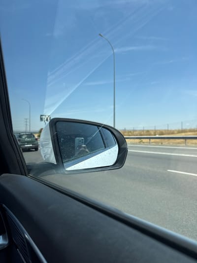 The image displays a daytime view from the interior of a vehicle, primarily focusing on the passenger-side window and external side mirror. The side mirror, which appears to be smudged or dirty, reflects a hand holding a light-colored smartphone with a triple camera array, indicating the photographer's presence and action. Beyond the vehicle, a multi-lane asphalt highway is visible, featuring white dashed lane markings. A dark green SUV is observed in the distance, traveling in the same direction or an adjacent lane. To the right of the highway, a metal guardrail runs parallel to the road, beyond which lies dry, tan-colored vegetation extending towards the horizon. A tall, curved grey street lamp pole stands prominently on the right side, with another similar pole visible in the far distance on the left. The sky is clear and bright blue, containing several faint white contrails, suggesting a fair weather environment. This scene is situated on a highway within or in the vicinity of San Sebastián de los Reyes, Spain.