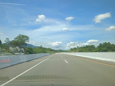 The image shows a straight asphalt highway, likely a toll road, situated in Seulimeum, Indonesia. The road is flanked by concrete barriers on both sides. Lush green vegetation and trees line the road, with hills visible in the distance. The sky is blue with scattered white clouds. There is a light-colored vehicle visible in the distance on the road. The photo appears to have been taken from inside a vehicle, as part of the dashboard is visible.