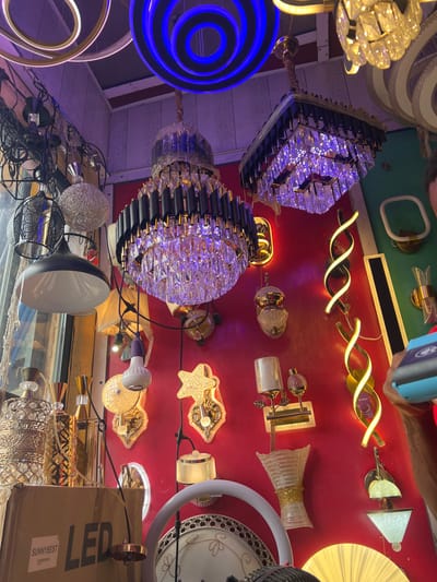 This image depicts the interior of a retail establishment specializing in lighting fixtures, located in Igbede, Nigeria. The scene is dominated by a dense display of various illuminated lights against red and green walls, and hanging from a light-colored wooden panel ceiling.
Suspended from the ceiling are multiple chandeliers, including two prominent multi-tiered designs featuring black and gold frames with clear crystal prisms, one of which is partially covered in protective plastic wrapping. Other hanging fixtures include circular LED rings, one emitting blue light and another gold, as well as metallic pendant lights, some with intricate patterns and a black dome-shaped light with a white interior.
The walls, primarily a vibrant red with a green section on the far right, are densely covered with a diverse array of wall-mounted lights. These include several illuminated spiral LED strips, star-shaped sconces, rectangular fixtures, and various decorative lamps with glass or metallic shades.
In the lower left, a brown cardboard box labeled "SUNNYBEST" and "LED" is visible. Partially visible in the lower right is a white circular ring light stand. The upper right corner reveals the shoulder and arm of an individual, possibly male, holding a light blue and black digital camera or smartphone, pointed towards the lighting display, suggesting the capture of the image itself. The overall environment is brightly lit by the numerous display items.
