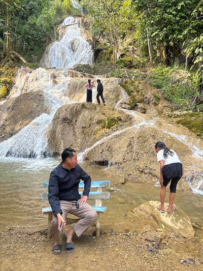 A man sits on a blue wooden bench, holding a cell phone, while a woman in a white shirt stretches on a rocky surface. Further in the background, two people stand by the cascades of a waterfall. The landscape is filled with lush greenery and rough terrain, suggesting an outdoor recreational area. The image is oriented sideways.