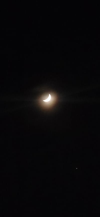 A low-angle, wide shot shows an apparent crescent moon against a black sky. The moon exhibits a bright, crescent shape with vertical diffraction spikes emanating from its lower portion. A small, indistinct star-like point of light is visible in the upper right of the frame. No people, subjects, objects, actions, interactions, or specific environmental features are visible other than the sky and celestial bodies. There is no information in the image to indicate the location is Los Alcázares, Spain.