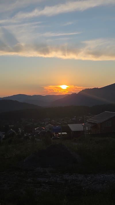 This image depicts a sunset in Ozernoe, Russia. The sun is low on the horizon, casting a warm, orange glow across the sky and silhouetting distant mountains. Wispy clouds are illuminated by the setting sun, creating a dramatic contrast with the clear blue sky to the left. In the foreground, several houses are visible, nestled amongst trees. The overall impression is one of a peaceful, rural evening. The time of day is clearly sunset, and the weather appears clear. No text is visible in the image.