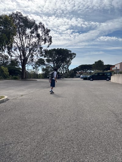 A medium shot, rotated counter-clockwise by 90 degrees, depicts a person skateboarding on an asphalt road in Fréjus, France. The skateboarder, facing towards the upper right of the frame, wears a white shirt, dark shorts, and a backpack. A skateboard is attached to the backpack. The sky, visible on the left, is light blue with scattered white clouds. Tall trees line the left side of the road. Parked vehicles are visible on the right side of the road next to a building. The road surface is asphalt.