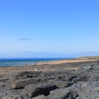 This image depicts a view of Fanore Beach in Fanore, Ireland, on a clear, sunny day. The foreground is dominated by large, dark, weathered rocks with uneven surfaces and some pools of water. Beyond the rocky shore, a sandy beach stretches out, populated by numerous people. Some individuals are walking along the shoreline, while others are in the shallow water, engaging in activities such as swimming or wading. Further out, the sea is a deep blue, with a line of distant land visible on the horizon, characterized by a range of hills dotted with wind turbines. The sky is a vibrant, cloudless blue.