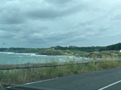 A cloudy day over the ocean. The water is a deep blue-green and choppy, with whitecaps. In the foreground, there is a wooden fence, and beyond it, green hills dotted with trees. The road is asphalt and runs along the coast. A white line is visible on the right side of the road. The scene appears to be a coastal highway, possibly in Bulgaria, given the location context.