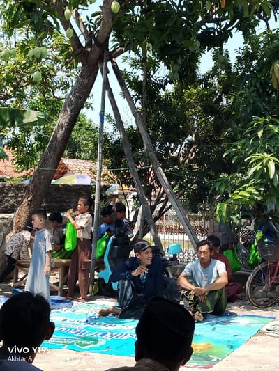 This image depicts an outdoor daytime gathering of approximately 10-15 individuals, predominantly males of varying ages from children to adults, in Jagapura Kulon, Indonesia. The scene is set under a large mango tree laden with unripe green fruit, its trunk supported by three substantial wooden poles tied to the main branches.
In the foreground, an adult male with his back to the viewer wears a black peci. On a blue and white patterned mat spread on the ground, two adult males sit cross-legged. The man on the left wears a black long-sleeved shirt and a traditional black head covering, smiling and looking to his right. Beside him, a man in a light grey t-shirt and a patterned sarong looks forward.
Behind these seated individuals, a light blue scooter is parked. To the left, several children and young boys are standing or leaning against a wooden bench; one boy in a light blue tunic stands upright, and another in a brown scout-like uniform holds a bright green bag. Additional children, some holding similar green bags or mobile phones, are partially visible in the mid-ground. On the far right, a red bicycle with a basket is present.
The background reveals residential structures with tiled roofs through dense green foliage. A white metal fence with decorative elements is also visible. The environment is brightly lit by direct sunlight, indicative of a clear day. The overall atmosphere suggests a casual community gathering.