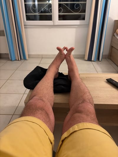 The image shows a person's legs from an overhead perspective. The person is wearing yellow shorts. Their lower legs are angled toward a black garment resting on a light brown wooden table. The person's feet are visible. In the background, there's a white wall with a window featuring decorative metal grilles. Above the window is a striped valance in shades of blue, white, and beige. The tile floor is beige. The scene's location is Fréjus, France.