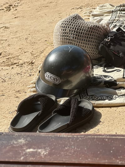 A helmet with a "Lucky" sticker sits on a sandy surface alongside a woven bag, a patterned cloth, and a black bag. Next to these items, a pair of black sandals labeled "PRIMARK" rest on the sand, partially obscured by a wooden post or wall on the right side. The items appear to be laid out in the sun, casting soft shadows on the textured ground.