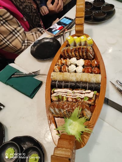 A sushi boat filled with a variety of sushi rolls including California rolls, spicy tuna rolls, and tempura rolls. The boat is wooden and shaped like a traditional Japanese vessel. Alongside the sushi is a plate with several small bowls containing soy sauce and wasabi. Cutlery wrapped in a green napkin is on the table. A cell phone is also on the table next to the boat.