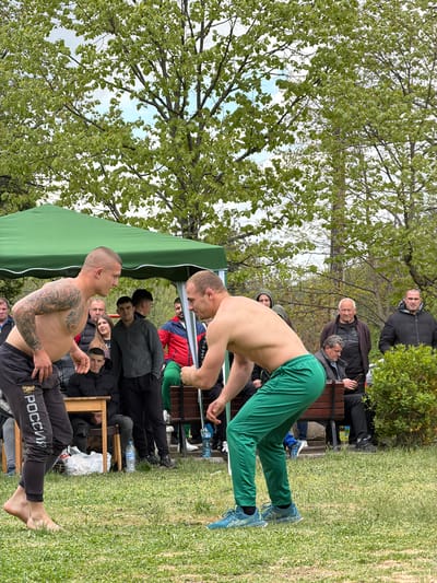 The image depicts two shirtless men engaged in a wrestling match outdoors, possibly a folk wrestling competition. One man is wearing green pants and blue athletic shoes, while the other wears black pants with "POCСИЯ" (Russia) printed on them and is barefoot. The match is taking place on a grassy area under a green canopy tent. Several spectators are seated and standing around the wrestling area. In the background are trees with green leaves. Based on the textual context given, this event is held in the city of Dulevo, Bulgaria.