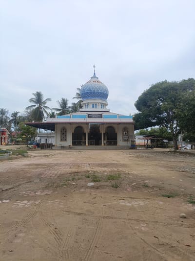 The image depicts the Meunasah Raya mosque in Meunasah Raya, Indonesia, under an overcast sky. The mosque features a prominent blue dome with intricate white patterns and a crescent moon finial. Its facade is white with blue trim and arched windows. A covered walkway extends from the mosque, supported by pillars. In the foreground, a dusty, unpaved area shows tire tracks and scattered debris. To the right of the mosque, a large tree shades a small shelter with a motorcycle parked underneath. Various structures, including palm trees and smaller buildings, are visible in the background, suggesting a somewhat rural or semi-developed setting. The overall scene is one of quiet midday stillness.