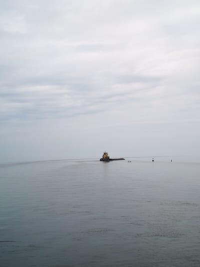 An overcast sky looms above a calm body of water, likely Lake Belyoye, near the village of Kurganovo, Russia. A tugboat, yellow and gray, is seen in the distance, moving away from the viewer. It is towing a long, dark barge. Two small figures appear to be on the barge. Further in the distance, two buoys mark the water. The water's surface exhibits gentle ripples, suggesting minimal wind. The overall scene is subdued, with muted colors due to the cloudy conditions.