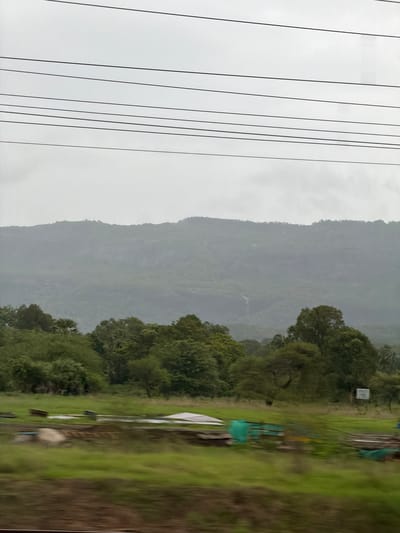 The image depicts a hazy, overcast sky above a lush green landscape. Multiple power lines stretch horizontally across the upper portion of the frame. Below the sky, a distant, mist-shrouded mountain range dominates the background, with faint indications of a waterfall or stream cascading down its slopes. In the mid-ground, a dense line of various trees forms a verdant barrier. The foreground reveals a field with patches of green grass, some debris including what appears to be plastic sheeting, metal structures, and a large rock. The scene suggests a rural or semi-rural environment, likely in the vicinity of Saphale, India, given the presence of monsoon-like weather conditions and the characteristic vegetation. There are no people visible in the image.