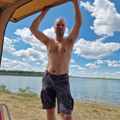 A shirtless man with a beard and tied-back hair, wearing dark cargo shorts and a belt, stands outdoors with his arms raised, holding onto the edge of a tan awning. He is positioned in the foreground, facing the camera. Behind him, a wide body of water, likely a lake, stretches to the horizon, bordered by a treeline. The sky is bright blue with scattered white clouds. The ground in front of the water is covered in dry grass and some sparse vegetation. Two fishing rods are set up on the shore in the mid-ground. The overall scene appears to be a recreational setting by a lake on a sunny day. This location is likely Gorni Dabnik, Bulgaria.
