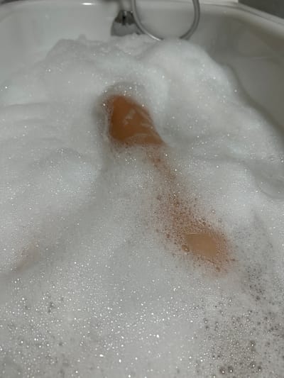 A person's leg is submerged in a bathtub filled with white bath foam. The leg appears to be light-skinned. The foam is bubbly and covers the majority of the image, obscuring the rest of the bathtub and any other objects within it. The scene is likely indoors, in a bathroom. There is no information in the image to suggest the location is in Vyshevychi, Ukraine.