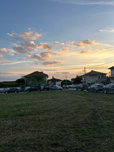 The image captures a street scene in Panicheri, Bulgaria, during what appears to be sunset. The sky is a mix of blue with scattered clouds tinged with orange and yellow hues, indicative of the setting sun. Several parked cars, including a dark blue hatchback, a black SUV, and other sedans and SUVs, are lined up along the side of a paved road. Behind the cars, a row of houses with tiled roofs is visible, interspersed with trees and vegetation. The foreground consists of a grassy area, appearing somewhat unkempt. Power lines and a street light are also present, suggesting a residential area. No people are visible in the frame.