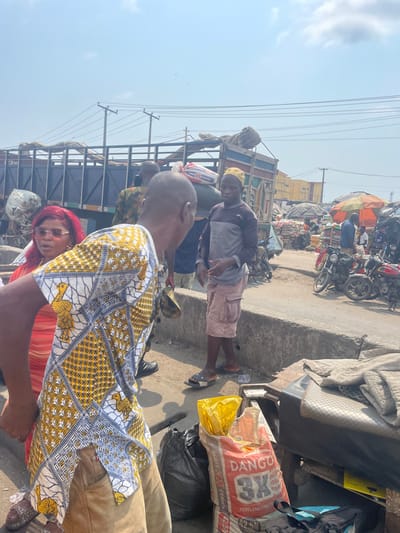 The image depicts a bustling outdoor market scene in Awjaw, Nigeria, during daylight hours under a clear to lightly clouded sky. In the foreground, a man with his back partially to the camera wears a short-sleeved patterned shirt featuring yellow, white, and blue geometric designs and stylized bird motifs, paired with light brown trousers. To his left, a woman with bright red hair and sunglasses wears a coral-colored top. In the mid-ground, a man with a light yellow skullcap stands on the far side of a concrete curb, wearing a two-tone long-sleeved shirt, beige cargo shorts, and black flip-flops.
The immediate foreground to the right contains a low, makeshift market stall topped with fabric or bedding, adjacent to several bags including a white sack labeled "DANGO 3X PORTLAND LIME CEMENT" with a yellow plastic bag inserted, and a black trash bag. A concrete curb separates the immediate foreground from a slightly lower ground level where the market extends.
In the background, a large blue flatbed truck, appearing heavily loaded with goods covered by tarps, is parked. Numerous individuals are visible around the truck and further into the market area. The market features multiple stalls, some covered with dark tarps and an orange umbrella, and stacks of goods in sacks. Several motorcycles are parked on the right side of the market. Overhead, power lines are visible against the sky. The ground throughout the scene appears dusty.
