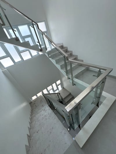 This is an interior, eye-level, medium shot of a modern, white, three-story staircase with a glass and stainless steel railing. The staircase has a landing between each floor, and each step is made of a light-colored stone with grey veining. The walls are painted white, and there are large windows on the second and third floors. The floor is tiled in a light grey. The scene is clean and well-lit, and the overall impression is one of spaciousness and elegance. There are no people in the image. This scene is not specifically identifiable as being in Tanjungmeru, Indonesia; it is a generic modern interior.