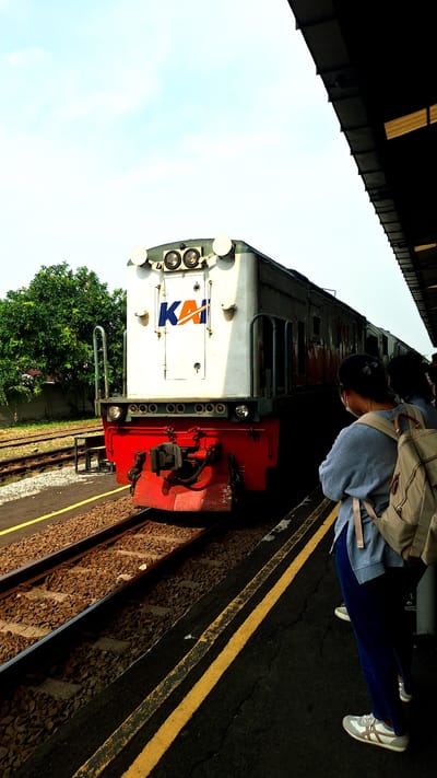 The image shows a train at a station in Karawang, Indonesia. A person wearing a gray sweatshirt, blue pants, and white sneakers stands on the platform. They have a backpack on. The train is predominantly white with a red front and the KAI (Kereta Api Indonesia) logo. The station platform has yellow lines marking the edge. Train tracks with gravel are visible beside the platform. Trees are visible beyond the tracks. The sky is clear and blue. The photo is taken at an angle.