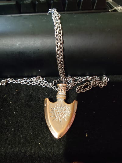 A copper-colored arrowhead pendant with intricate etchings, including a triangular symbol with radiating lines, hangs from a silver-toned metal chain. The pendant is positioned centrally in the frame, lying on a black, velvety surface. The chain is draped across the surface and extends upwards towards a dark, textured background, which appears to be the surface of a keyboard. The lighting is overhead and casts subtle shadows. There are no people or animals present. The scene is static and appears to be a product photograph or personal item display. The location is not discernible from the image itself.