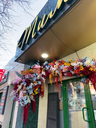 The image displays the exterior of a building, viewed from a low angle looking upwards, likely a commercial establishment located in Ol'ginskaya, Russia. The most prominent feature is an elaborate and colorful floral arrangement adorning the facade above the entrance.
This vibrant floral display is composed of a multitude of artificial flowers and foliage in various shades, including bright reds, deep blues, soft pinks, sunny yellows, whites, purples, and oranges. Long, slender red and yellow decorative elements extend from the main floral mass. A single circular light fixture is mounted on the golden-brown facade, illuminating the area beneath the floral garland. A small security camera is also visible near the light.
The building's exterior features a smooth, reflective golden-brown panel at the top, below which is a dark green, possibly marble, section. A lighter beige or cream-colored section is also visible, with what appear to be light green tiles or panels. A green-framed glass door and window are present on the right side of the image, through which the interior of a shop can be faintly discerned. Reflections in the glass show elements from the opposite side of the street.
Visible text includes Cyrillic script on the green door, which transliterates to "thyme flowers" and "@thyme_flowers", along with "круглосуточно" (kruglosutochno, meaning 24/7 or around the clock), strongly suggesting a flower shop with extended hours. Another red sign at the bottom right reads "ТОЧКА" (Tochka, meaning point) and "ЗАТОЧКА" (Zatochka, meaning sharpening), possibly indicating a service point. Other partially visible text from OCR includes "В ПРОДАЖЕ" (V Prodazhe, meaning "for sale") and "SERVICE PROFI". On the golden-brown facade, large, stylized golden letters are partially visible, reflecting the surrounding environment.
The sky, visible through the bare branches of a tree on the left, appears bright, indicating daytime. The absence of leaves on the tree suggests it might be late autumn or winter. No people are present in the image.