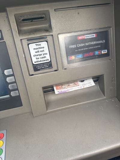 The image shows a close-up view of an ATM in Welshpool, United Kingdom. The ATM displays signage indicating "FREE CASH WITHDRAWALS" and "This machine will not charge you for cash withdrawals." Bank cards from MasterCard, Cirrus, LINK, Maestro, and VISA are accepted. A ten-pound note is partially dispensed from the cash outlet. A customer assistance phone number is listed on the ATM.