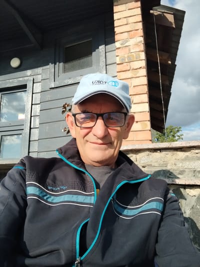 A man wearing a light blue baseball cap and glasses poses for a selfie outdoors. He is wearing a dark jacket with light blue and white stripes. The background shows a dark grey wooden building with windows, a brick chimney, and a stone wall. The sky is partly cloudy. The man appears to be standing or sitting outdoors, possibly in front of a residential structure.