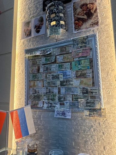 The image depicts a wall decorated with photographs and framed currency, seemingly a collection of mementos. A large framed display contains numerous banknotes, many of which have handwritten messages and dates. Above this frame, two smaller photographs are affixed to the wall, showing groups of people. To the right of these, a larger holiday-themed postcard features three individuals and animals in a snowy setting. The wall itself has a textured, light-colored finish. In the foreground, partially visible at the bottom of the image, are two flags, one appearing to be the Russian flag, and several glass containers, possibly salt and pepper shakers.
The presence of the Russian flag and the Cyrillic script on the banknotes and photographs strongly suggests a connection to Russia. Suifenhe, China, is a border city with Russia, known for its trade and tourist activity involving Russian citizens. This context suggests the location might be a business or establishment frequented by Russians, where they leave these monetary tokens as souvenirs or expressions of gratitude. The style of the decorations and the personal inscriptions point towards a casual, communal space where patrons might leave their mark.