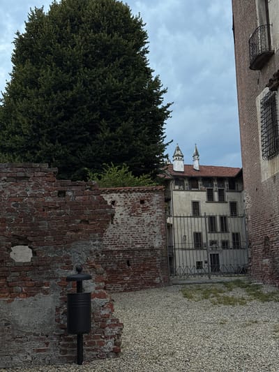 A wide shot captures a courtyard in Lagnasco, Italy, bordered by an old brick wall on the left and a tall brick building on the right. A large, dense green tree stands prominently on the left, partially obscuring a white building with a red-tiled roof visible in the distance. This building features arched windows and two distinctive white spires. A wrought-iron gate separates the courtyard from the distant building, and a black trash receptacle is situated against the brick wall in the foreground. The ground is covered in a layer of small, light-colored stones with sparse patches of green grass. The sky is overcast, with a mixture of gray clouds.