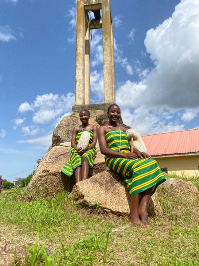 In Dutsen Kato, Nigeria, two young girls are captured in a posed photograph against a backdrop of a bright blue sky with scattered white clouds. The image is oriented vertically, with the subjects positioned on a large, weathered rock formation. Both girls are dressed in vibrant, traditional attire featuring green and yellow stripes with thin red accents. The fabric has visible text indicating it was "PRINTED BY SUPER MK.K."
The girl in the foreground, seated higher on the rock, is looking towards the camera with a warm, engaging smile. She wears a necklace and holds a fluffy, white object in her hands, possibly part of a costume or cultural accessory. Her legs are crossed, and her bare feet rest on the rock. The second girl is positioned slightly behind and below her, her face partially obscured but also appearing to be engaged with the camera. She also wears the same striped outfit and holds a similar white accessory.
To the left of the frame, a large concrete structure with visible rebar rises towards the sky, suggesting the presence of a building or tower nearby. A portion of a red-roofed building is visible in the upper right of the image, beyond the rock formation. The ground around the rock is covered in dry grass, indicating a potentially warm or dry climate. The lighting suggests it is daytime, with the sun casting subtle shadows. The overall mood of the image appears cheerful and celebratory, possibly capturing a cultural event or a moment of joy.