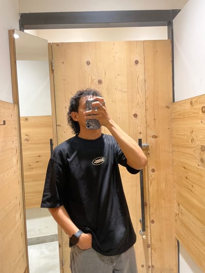 A person is taking a mirror selfie in a fitting room. The person is wearing a black t-shirt with a white oval logo and grey pants. They are also wearing rings on their fingers and a smartwatch on their left wrist. The fitting room has wooden walls and a mirror. The image is taken from a medium shot, focusing on the person and their reflection. The background shows the interior of what appears to be a retail establishment, possibly in a mall or clothing store located in Kelapa Dua, Indonesia. The lighting is adequate for the photograph.