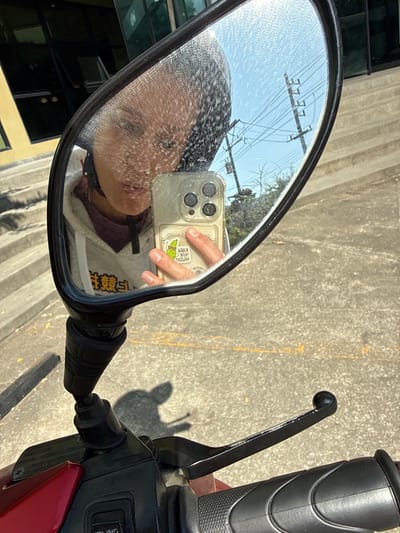 A selfie is reflected in a motorcycle mirror. The mirror is slightly dirty and shows the person holding a smartphone, presumably taking the photo. The motorcycle handle and levers are visible, casting a shadow on the concrete surface. Overhead wires are also visible in the mirror. The person's reflection shows a masked face and part of a patterned sweatshirt.
