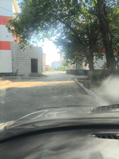 The image shows a view from inside a vehicle looking out onto a dirt road in the city of Abay, Kazakhstan. The road is lined with trees and buildings. On the left, there is a white building with orange accents and a section of light-colored brick wall with a dark door and steps. On the right, there are more trees and a white building with windows. In the distance, there are other buildings visible, and two white cars are parked on the side of the road. The sun is shining, casting shadows on the road. The foreground shows the dashboard of the vehicle.