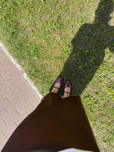 The image shows a pair of burgundy flat shoes with ankle straps and a portion of a dark brown skirt. The footwear and clothing are positioned on a grassy area next to a brick pathway. A shadow, likely cast by the person wearing the clothing and shoes, stretches across the grass. The scene appears to be outdoors during the daytime, with ample sunlight creating distinct shadows. The environment consists of green grass and a brick-paved walkway, suggesting an urban or suburban park or sidewalk. The location is not specified in the image, and there is no information provided about Barysaw, Belarus.