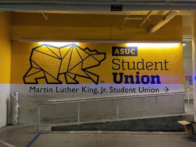 A large sign on a yellow wall with a black geometric bear graphic and "ASUC Student Union" text is displayed above "Martin Luther King, Jr. Student Union" with an arrow pointing right. The sign is illuminated by a bright light above. Below the sign, a concrete ramp with metal railings leads upwards. A blue line is painted on the floor, and a cardboard box sits to the right. The location appears to be an indoor parking garage.